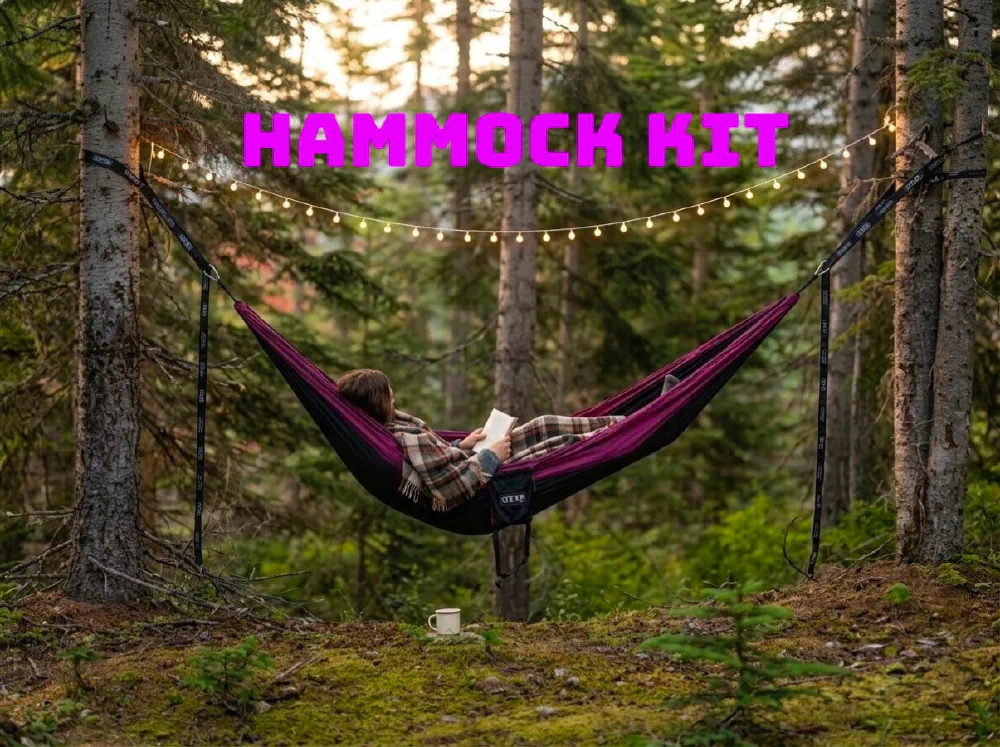 A person reading in a purple camping hammock strung between two conifers, with warm string lights hung above and a mug on the moss below