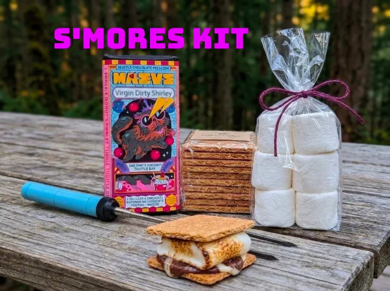 S'mores kit on a picnic table: a Maeve bar from Seattle Chocolate, graham crackers, a bag of marshmallows, roasting sticks and an assembled s'more