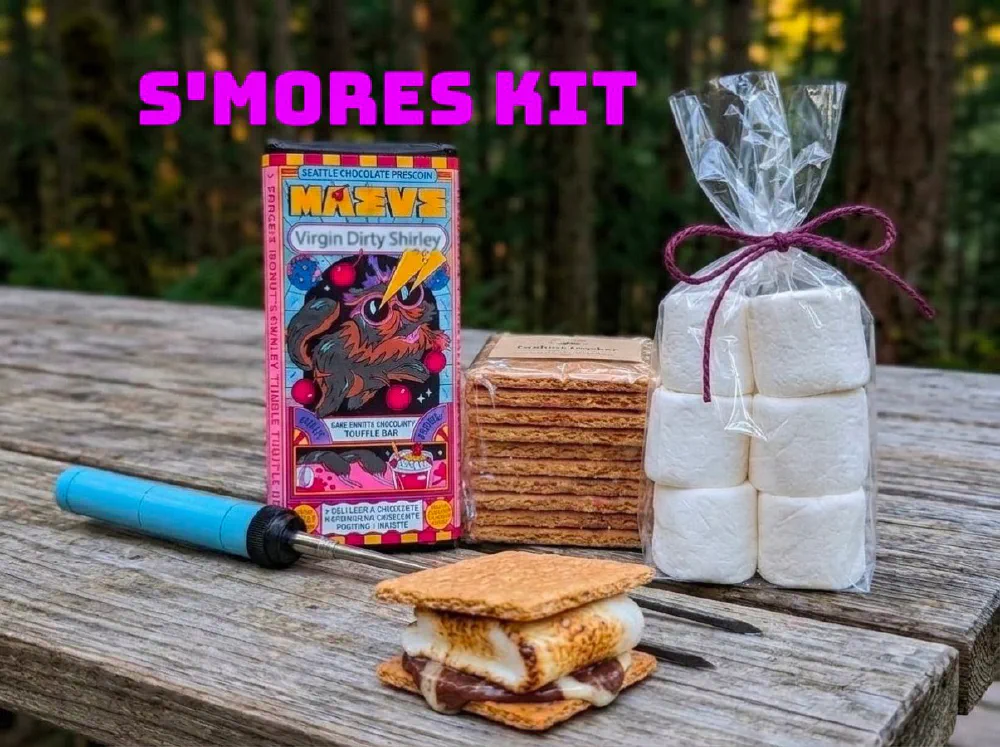 S'mores kit on a picnic table: a Maeve bar from Seattle Chocolate, graham crackers, a bag of marshmallows, roasting sticks and an assembled s'more